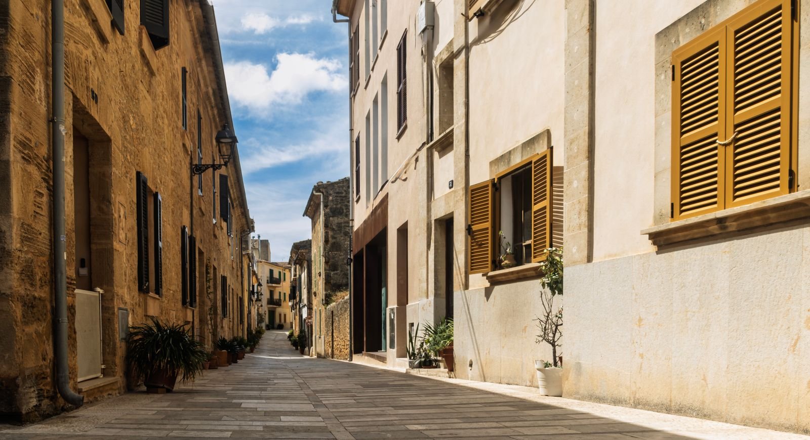 Ruelle méditerranéenne aux façades claires et aux volets fermés, avec l'ombre portée franche du plein soleil